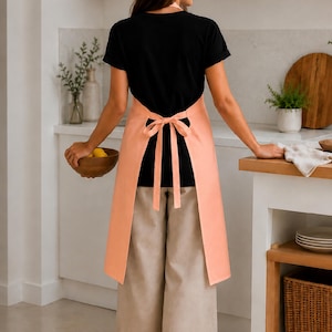Könnte beinhalten: Eine einfarbige pfirsichfarbene Schürze mit Bindegürtel in der Taille, getragen über einem schwarzen kurzärmeligen T-Shirt und einer weit geschnittenen beigen Hose. Die Schürze bedeckt Rücken und Seiten und wird mit einer Schleife im Rücken gebunden.