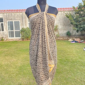 May include: A sarong featuring a dark brown and cream geometric pattern with a repeating floral-like motif. The fabric is draped around a mannequin in a halter style, showcasing a solid cream border along the edges and a soft, flowing silhouette.
