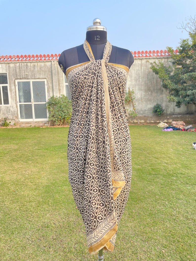 May include: A sarong featuring a dark brown and cream geometric pattern with a repeating floral-like motif. The fabric is draped around a mannequin in a halter style, showcasing a solid cream border along the edges and a soft, flowing silhouette.