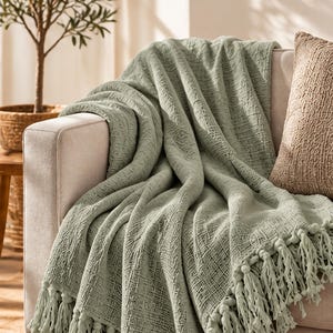 Può includere: Una coperta color verde salvia con un tessuto testurizzato a trama aperta e lunghe frange annodate lungo il bordo inferiore. La coperta è drappeggiata sul bracciolo e sulla seduta di un divano di colore chiaro, evidenziando la trama tessuta.