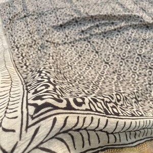 May include: A cream-coloured textile featuring a dark brown block-printed pattern of repeating circular and geometric shapes. The fabric is bordered by a dark brown leaf-like motif and finished with a metallic gold band along the edge.