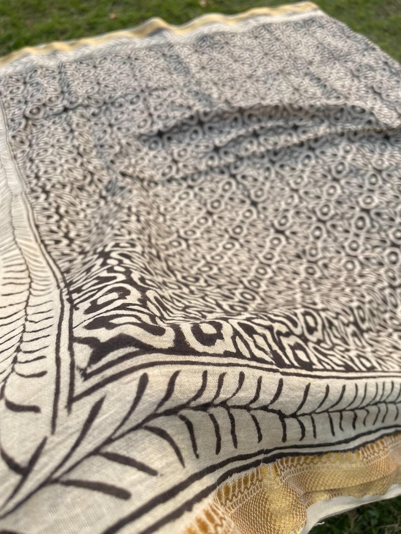 May include: A cream-coloured textile featuring a dark brown block-printed pattern of repeating circular and geometric shapes. The fabric is bordered by a dark brown leaf-like motif and finished with a metallic gold band along the edge.