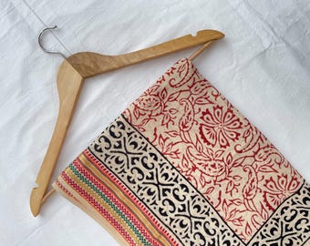 May include: A light beige fabric textile draped over a wooden hanger. The fabric features a red floral paisley pattern, a wide black geometric border, and a thin decorative stripe with green, red, and yellow accents along the bottom edge.