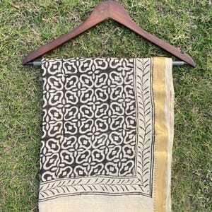 May include: A rectangular textile piece featuring a dark brown geometric pattern on a cream background, framed by a leaf-like border and a solid gold-toned edge. The fabric is draped over a dark wooden clothes hanger with a silver metal hook.