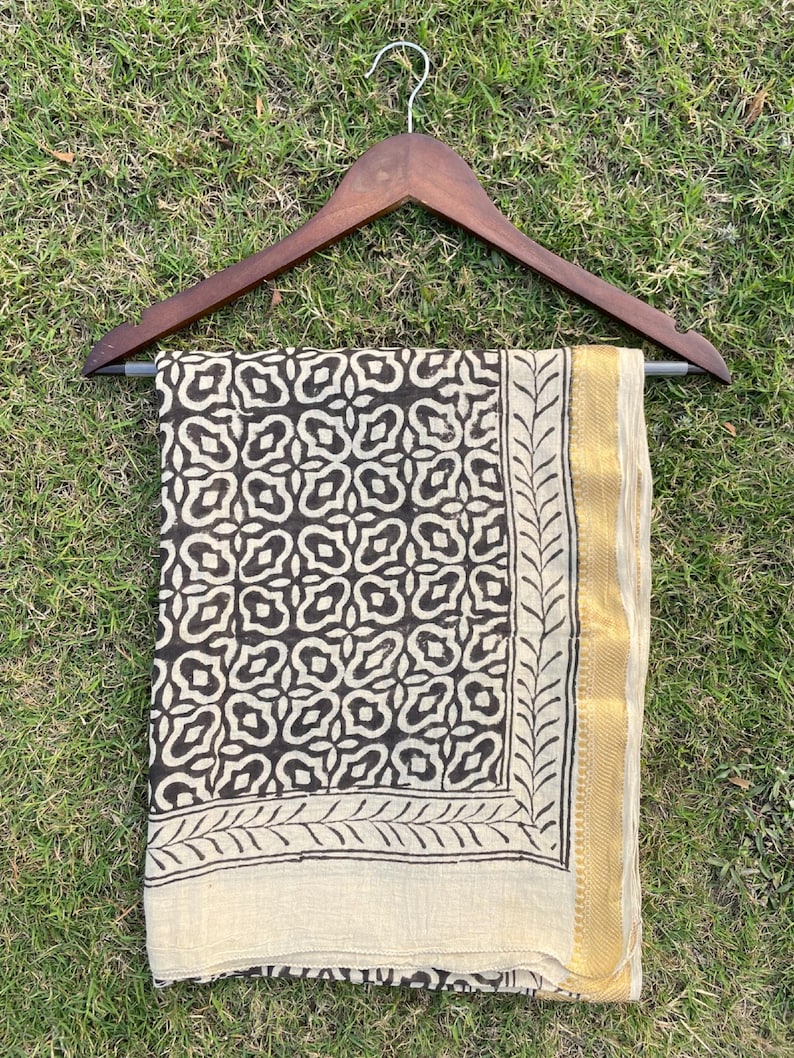 May include: A rectangular textile piece featuring a dark brown geometric pattern on a cream background, framed by a leaf-like border and a solid gold-toned edge. The fabric is draped over a dark wooden clothes hanger with a silver metal hook.