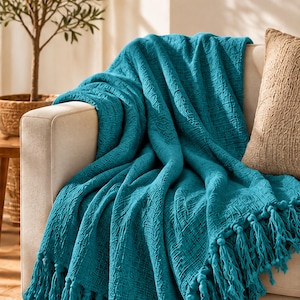 May include: A teal throw blanket with a textured, woven fabric construction and long, knotted fringe tassels along the bottom edge. The blanket is draped over the arm and seat of a light-colored sofa, showcasing its deep color and intricate weave.