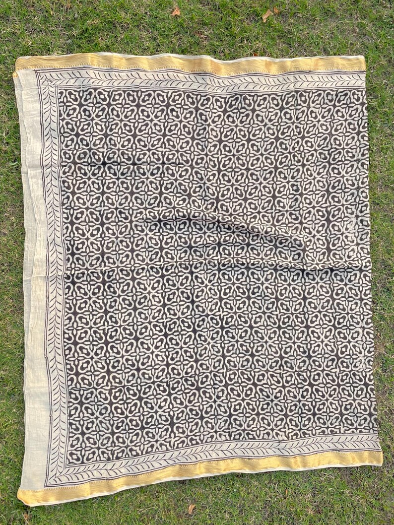 May include: A rectangular textile featuring a dark brown repeating geometric pattern on a cream background. The fabric is bordered by a thin, dark brown line and a wider, solid gold-coloured edge, creating a distinct frame around the central design.