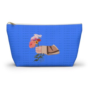 May include: A blue zippered pouch featuring a repeating pattern of horizontal dashed lines. Centered on the front is a graphic illustration of a hand wearing a tan sleeve, holding a colorful bouquet of pink, orange, and yellow flowers with green stems.