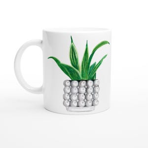Könnte beinhalten: Ein weißer Keramikbecher mit einer zentrierten Illustration einer grünen Bogenhanfpflanze mit spitzen Blättern. Die Pflanze steht in einem strukturierten, hellgrauen Topf, der mit einem Muster aus kleinen, runden Kugeln verziert ist.
