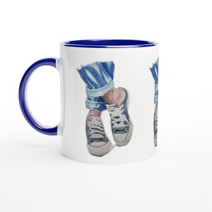 May include: A white ceramic mug with a dark blue handle and interior rim. The exterior features a repeating illustration of blue denim jeans with rolled cuffs and low-top white canvas sneakers with blue laces and white rubber toe caps.