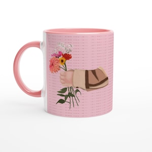May include: A pink ceramic mug featuring a graphic illustration of a hand holding a bouquet of colorful flowers including pink, orange, and yellow blossoms with green stems. The mug has a solid pink handle and a light pink background with subtle lines.