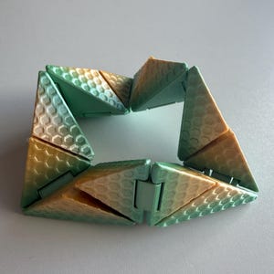 May include: A geometric bracelet made of linked triangular sections. The sections have a honeycomb pattern and a colour gradient, shifting from green to gold. The bracelet is designed to fold and unfold.