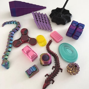 May include: A selection of colourful fidget toys in different shapes and sizes. Includes a purple segmented snake, a red and black fidget spinner, a pink push button, a red and brown dragon, and a black spinning top. The toys are arranged on a white surface.