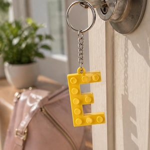 May include: A bright yellow letter 'E' keyring, constructed from interlocking blocks, suspended from a silver chain and key ring. The keyring is attached to a door lock. A pink handbag and a potted plant are in the background.