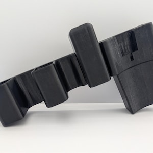Pode incluir: Uma escultura abstrata preta com um design geométrico. A escultura apresenta uma série de blocos retangulares e bordas curvas. O objeto é feito de um material escuro e sólido e está sobre um fundo branco.