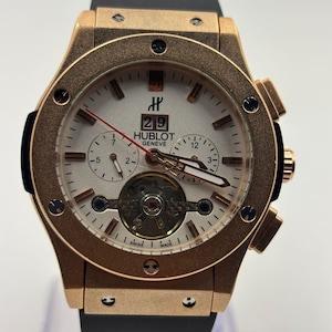 May include: A gold-toned Hublot Geneve watch with a white face, black band, and red second hand. The watch features a date display, sub-dials, and visible internal mechanisms. The watch is displayed on a clear stand.