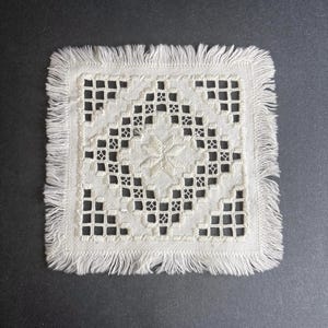 May include: Square, off-white coaster with a detailed embroidered design. The center features a star-like pattern, surrounded by a geometric diamond and square motif. The edges are finished with a fringe.