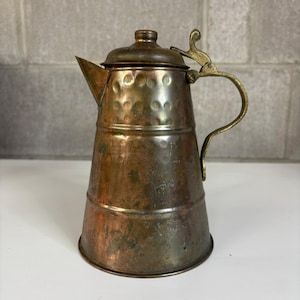 May include: A vintage copper teapot with a brass handle and lid. The teapot has a conical shape with a spout and a decorative handle. The copper has a patinaed finish, giving it an antique look. The lid is secured with a small knob.