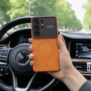 Może przedstawiać: Pomarańczowe etui na telefon z czarnym modułem aparatu, trzymane przed kierownicą samochodu. Etui ma okrągły wzór z tyłu i napis "4K 120 FPS".