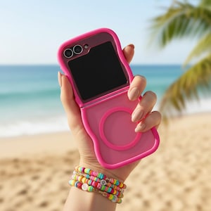 Może przedstawiać: Jasnoróżowe etui na telefon, otwarte, aby odsłonić czarny ekran, na tle plaży. Etui ma okrągły element konstrukcyjny i ochronny zderzak. Na nadgarstku znajduje się stos kolorowych bransoletek z koralików.