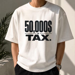 Pu&ograve; includere: Una t-shirt bianca a maniche corte con un testo nero in grassetto sul petto che recita 50,000$ sopra una linea orizzontale, con la parola TAX. stampata in grandi lettere maiuscole nere sotto la linea, incluso un piccolo simbolo pi&ugrave;.