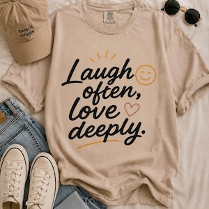 May include: Beige t-shirt with the text "Laugh often, love deeply" in black script, a smiling face, and a heart. The image also includes a beige baseball cap, sunglasses, blue jeans, and white trainers.