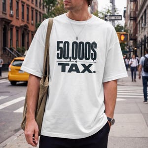 Pu&ograve; includere: Una t-shirt bianca a maniche corte con la scritta nera 50,000$ + TAX. stampata sul petto. Il capo presenta un girocollo e una vestibilit&agrave; ampia. Una borsa tote in tela marrone &egrave; portata a spalla insieme a una sottile collana a catena.
