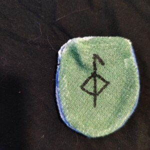 May include: A green patch with a blue border and a black symbol. The patch is a rounded rectangle shape. The symbol is a vertical line with a diamond shape in the middle and a line extending upwards.