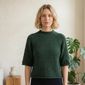 Könnte beinhalten: Ein waldgrüner Strickpullover mit kurzen Ärmeln und hohem Ausschnitt. Der Pullover besteht aus einem strukturierten Material und wird von einer Person getragen. Der Hintergrund umfasst eine Pflanze und einen kleinen Holztisch.