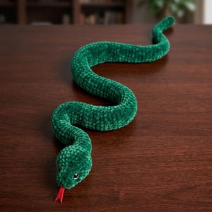 Op de afbeelding: Een groene, pluche slangenspeelgoed met een rode tong, liggend op een donker houten oppervlak. De slang is gemaakt van een zacht, getextureerd materiaal en het lichaam is gebogen in een kronkelend patroon. De kop van de slang heeft kleine, zwarte ogen.