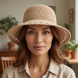 May include: A beige crochet hat with a rounded crown and a wide brim. The brim features a decorative openwork pattern along the edge, constructed from a textured, woven material. The hat has a structured shape and a neutral, light tan color.