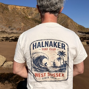 Może przedstawiać: Kremowy t-shirt z krótkim rękawem z grafiką na plecach. Wzór przedstawia dużą falę oceaniczną, surfera, palmy i wiatrak. Napis głosi HALNAKER SURF CLUB, WEST SUSSEX oraz SINCE 1954, wykonany niebieskim i czerwonym tuszem.