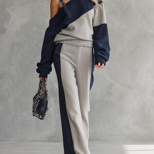 May include: A two-piece lounge set consisting of a long-sleeved, off-the-shoulder top and wide-leg trousers. The set features a colour-blocked design in navy blue and light grey. The top has a single shoulder strap, and the trousers have an elastic waistband.