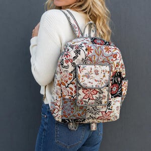 Könnte beinhalten: Ein gesteppter Rucksack mit Patchwork-Design und Blumenmustern in Rot-, Blau-, Schwarz- und Cremetönen. Die Tasche verfügt über eine Vordertasche mit Klappenverschluss, verstellbare Schultergurte und schwarz-weiß gestreifte Zierleisten.