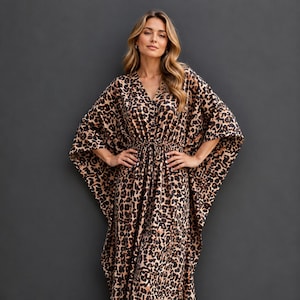 Op de afbeelding: Een enkellange kaftan-jurk met een bruin en zwart luipaardpatroon, een V-hals, wijde vleermuismouwen en een getailleerde taille met een trekkoord. Het kledingstuk wordt gecombineerd met bruine leren slippers met gekruiste bandjes.