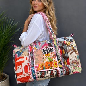 May include: A large rectangular duffel bag made from quilted fabric featuring a vibrant patchwork design. The exterior displays various patterns including floral motifs, geometric shapes, and architectural elements in red, yellow, blue, and black.