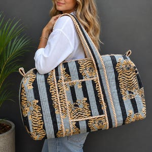 Könnte beinhalten: Eine große, rechteckige, gesteppte Reisetasche mit einem sich wiederholenden Tigermuster auf einem Hintergrund aus hellblauen und schwarzen vertikalen Streifen. Die Tasche hat dicke Stoffgriffe und eine Vordertasche mit heller Flechtkante.