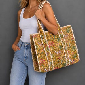 May include: A large rectangular tote bag featuring a vibrant mustard yellow base with a dense floral and botanical print in shades of pink, green, and white. The bag has structured sides and sturdy handles with a white and tan striped trim.