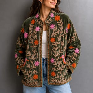 Op de afbeelding: Een olijfgroen fluwelen jasje met een opstaande kraag en een open voorzijde. Het jasje is versierd met ingewikkeld borduurwerk van roze en oranje bloemen, bladerrijke ranken en kleine vogelmotieven op de voorpanden en de mouwen.