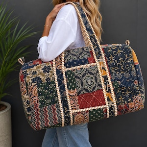Puede incluir: Una bolsa de viaje rectangular grande con un diseño de patchwork multicolor que presenta varios patrones florales y geométricos en tonos azul marino, rojo, verde y amarillo. La bolsa tiene bordes gruesos cosidos en color crema y dos asas.
