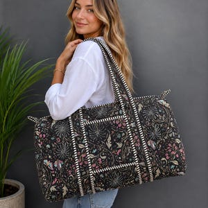 Könnte beinhalten: Eine große, rechteckige, gesteppte Reisetasche mit schwarzem Grund und einem detaillierten Blumen- und Schmetterlingsmuster in Rosa-, Blau- und Cremetönen. Die Tasche verfügt über dicke, schwarz-weiß gestreifte Stoffriemen und Besätze.