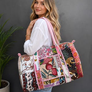 Può includere: Una grande borsa da viaggio rettangolare trapuntata con un vivace design patchwork composto da vari motivi floreali, botanici e animali nei toni del rosa, rosso, verde e nero, rifinita con bordi in tessuto a righe rosa e bianche e due tracolle.