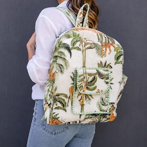 May include: A quilted fabric backpack featuring a cream-coloured base with a green and orange jungle-themed print of giraffes, leopards, and palm leaves. The bag includes a front pocket with green and white striped trim and matching shoulder straps.