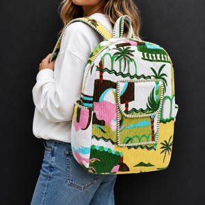 Könnte beinhalten: Ein Rucksack aus Kord mit einem lebendigen, mehrfarbigen tropischen Landschaftsdruck mit Palmen, Hügeln und abstrakten Formen in Gelb, Grün, Rosa und Weiß. Die Tasche verfügt über eine Reißverschlusstasche auf der Vorderseite und gepolsterte Träger.