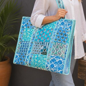 Könnte beinhalten: Eine große rechteckige Tragetasche mit einem Patchwork-Design aus verschiedenen blau-weißen Blumen- und geometrischen Mustern. Die Tasche hat zwei Schulterriemen und eine gesteppte Textur, die eine lebendige Mischung aus Azurblau, Türkis und Marineblau zeigt.