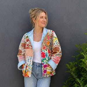 Könnte beinhalten: Eine gesteppte Jacke mit einem lebendigen Patchwork aus Blumenmustern in Rot-, Orange-, Gelb- und Grüntönen. Die Jacke hat einen hellblau-weiß gestreiften Kragen und Ärmelbündchen, ein weißes Tanktop, eine hellblaue Jeans und beigefarbene Schuhe.