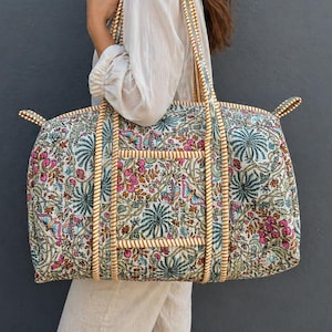 Könnte beinhalten: Eine große, gesteppte Reisetasche mit einem bunten Blumen- und Schmetterlingsmuster in Rosa-, Grün- und Blautönen. Die Tasche hat eine strukturierte, rechteckige Form mit dicken, cremefarbenen und beigen Streifengriffen und passender Kantenverzierung.