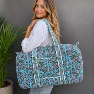 Könnte beinhalten: Eine große, rechteckige gesteppte Reisetasche mit hellblauem Grund und einem sich wiederholenden Blumenmuster in Rosa-, Grün- und Dunkelbrauntönen. Die Tasche verfügt über zwei stabile Schulterriemen und eine weiß-grün gestreifte Kante.
