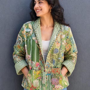 May include: A green floral patterned jacket with a striped collar and cuffs. The jacket features a patchwork design with various floral and leaf patterns in shades of green, red, and yellow. The jacket has two pockets and is worn over a white top and blue jeans.