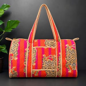 Könnte beinhalten: Eine gesteppte Reisetasche mit einem lebendigen orange-pinken vertikalen Streifenmuster und wiederkehrenden Tigerillustrationen. Die Tasche verfügt über zwei lange Tragegriffe, einen Reißverschluss und eine weiß-orange geflochtene Kante.